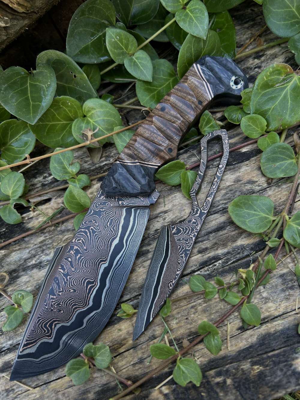 A unique knife with a brown and black crafted handle and a blade with a copper, black, and silver pattern.